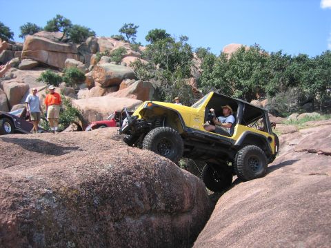 2001 Jeep Wrangler - Yellow Jeep Wrangler rock crawling over large granite boulders at an off-road trail event, with spectators in orange shirts watching.