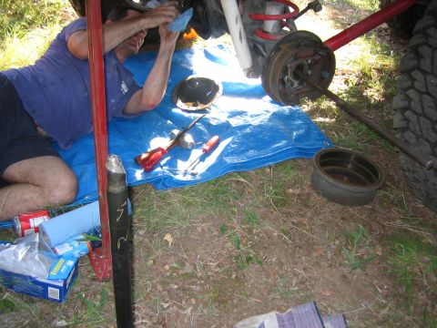2001 Jeep Wrangler - Outdoor trailside brake job: a person kneels on a blue tarp working under a lifted vehicle while the rear drum brake sits fully exposed, ba…