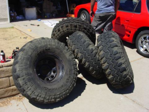 1994 Ford Bronco - A collection of four aggressive off-road tires are displayed on a driveway, with a red vehicle partially visible in the background.