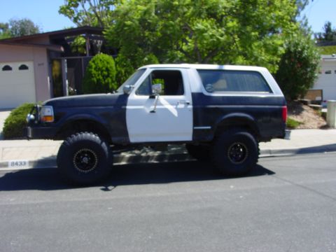 1994 Ford Bronco - Side view of a lifted dark blue Ford Bronco parked on a suburban street, wearing a white removable hardtop and a mismatched white driver's…