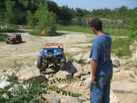 1992 Ford Bronco - Two off-road vehicles navigate a rocky terrain near a body of water under a clear sky.