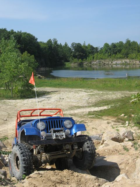 1992 Ford Bronco - A blue off-road jeep navigates a rocky, sandy path beside a calm lake under a clear sky.