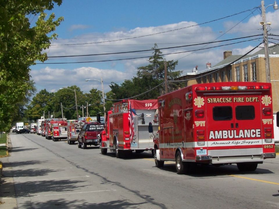 Red ambulance with 'SYRACUSE FIRE DEPT. AMBULANCE' on rear, in procession with fire trucks