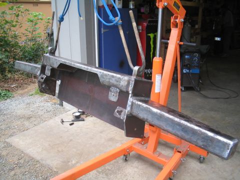 1979 Ford Bronco - A large shop-built bare-steel truck bumper hangs from an orange hydraulic engine hoist by blue rope slings at a garage doorway, its wing en…