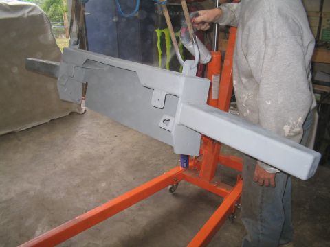 1979 Ford Bronco - A gray bumper part is being worked on, suspended by an orange engine hoist in a workshop.