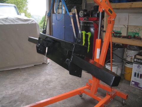 1979 Ford Bronco - A shop-built black steel front bumper hanging from blue straps on an orange folding engine hoist in a garage, showing its boxed tube face,…
