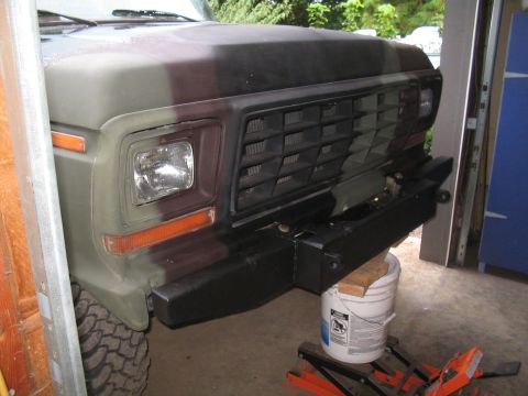 1979 Ford Bronco - Front three-quarter view of a lifted Ford truck painted in flat green-and-brown camo, sitting in a garage doorway while a shop-built black…