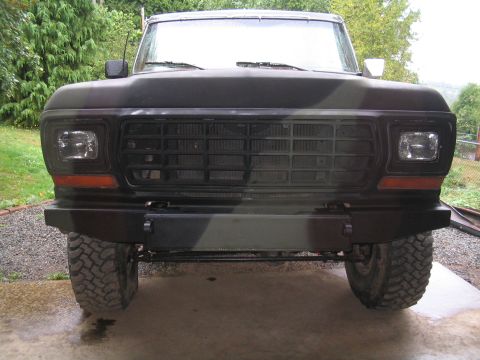 1979 Ford Bronco - Straight-on front view of a lifted Ford truck in flat camo paint with a white roof, parked on a wet concrete pad with grass and evergreens…