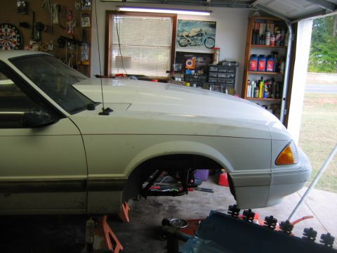 1992 Ford Mustang - Front end of a white coupe is disassembled while on jack stands in a garage.