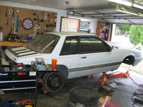 1992 Ford Mustang - A white 1992 Ford Mustang coupe is elevated on jack stands in a garage, undergoing maintenance.