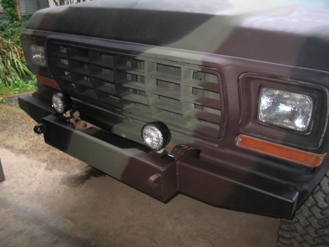 1979 Ford Bronco - Close-up of the passenger-side front corner of a Ford truck painted in flat camouflage green, brown and black, showing the egg-crate grille…
