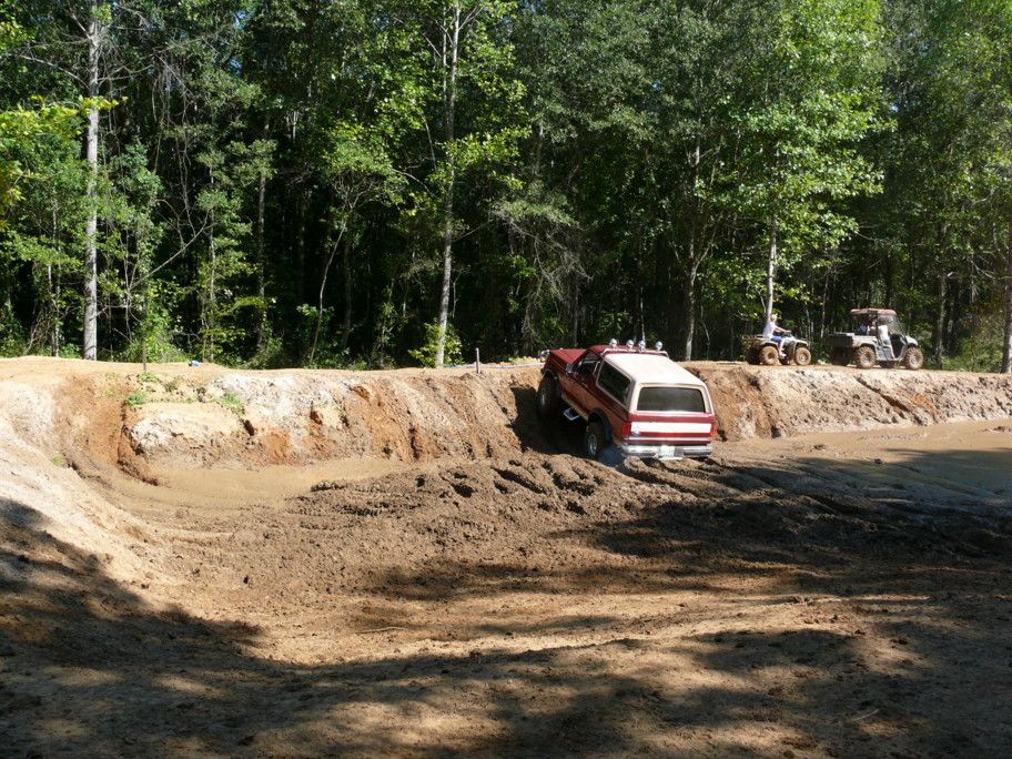1988 Ford Bronco climbing out of a mud pit, two ATVs on the bank behind it