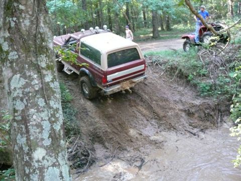 1988 Ford Bronco - Two off-road vehicles navigate a challenging muddy trail in a wooded area.