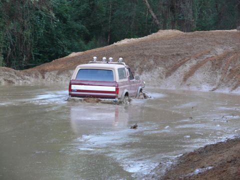 1988 Ford Bronco - A Ford Bronco navigates through a water-filled mud pit, kicking up water and mud.