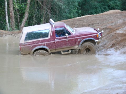 1988 Ford Bronco - A maroon Ford Bronco with a tan top is stuck in a muddy puddle, with trees in the background.