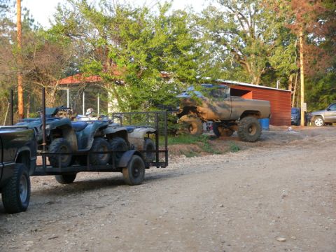1988 Ford Bronco - Pickup truck hauls ATVs on a trailer, while a lifted truck sits parked off-road.