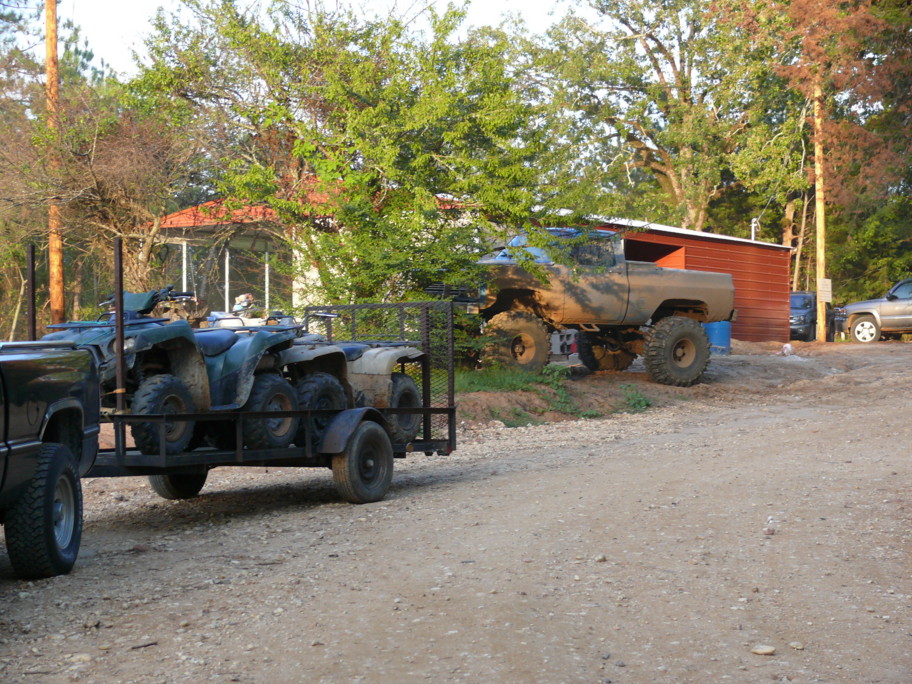 Pickup truck towing a trailer loaded with four ATVs, with a lifted mud-covered truck parked nearby