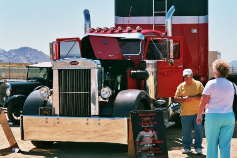1981 Kenworth W900A - A classic Peterbilt semi-truck is displayed with a vintage car in the background. A sign provides event details.