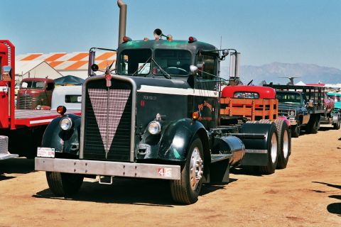 1981 Kenworth W900A - A line of trucks, including a dark green semi-truck, are parked outdoors under a clear blue sky.