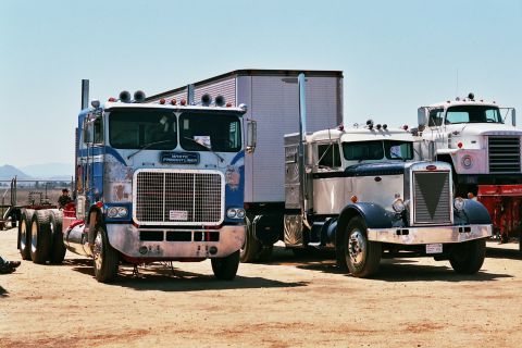 1981 Kenworth W900A - Two semi-trucks, one a White Freightliner, are parked on a dirt lot under a clear blue sky.