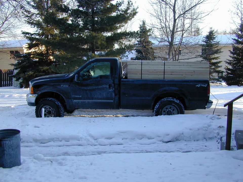 Side view of dark Ford F-350 4x4 regular cab pickup hauling stacked lumber in snowy driveway
