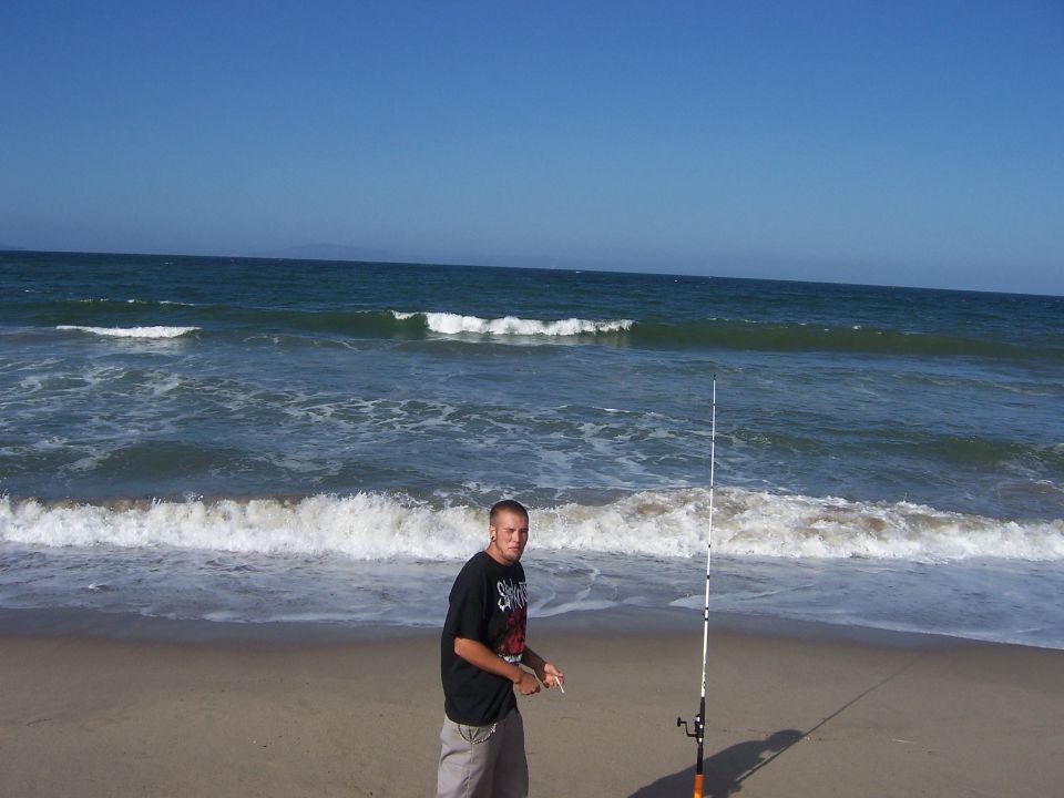 Guy holding a cigarette and fishing pole on a sandy beach with waves crashing