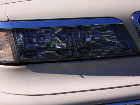 1996 Mercury Grand Marquis - Close-up of the driver-side headlight assembly on a light cream/tan sedan, showing the clear multi-reflector lens with amber turn-signal se…