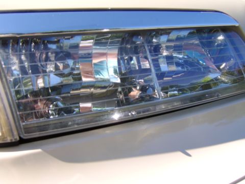 1996 Mercury Grand Marquis - Close-up of a headlight assembly on a light silver/white vehicle, showing the clear lens with multiple reflector segments, chrome trim, and…