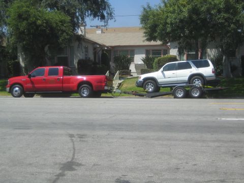 1968 Ford Mustang - A red pickup truck is seen towing a white SUV on a trailer down a residential street.