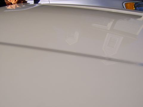 1996 Mercury Grand Marquis - Close-up of a glossy white body panel, likely a hood or trunk lid, showing a mirror-like reflection of a house with windows and an amber co…
