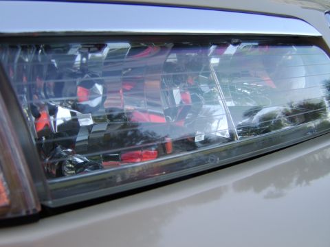 1996 Mercury Grand Marquis - Close-up of a headlight assembly on a light silver/beige vehicle, showing the clear lens with visible multi-reflector optics, chrome intern…