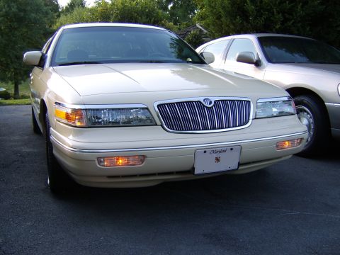 1996 Mercury Grand Marquis - A cream/beige Mercury Grand Marquis sedan photographed from the front, showing its chrome vertical-bar grille, dual headlights, amber turn…