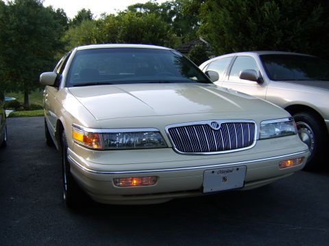 1996 Mercury Grand Marquis - A cream/beige Mercury Grand Marquis sedan photographed from the front three-quarter angle, showing its chrome waterfall grille with the Mer…