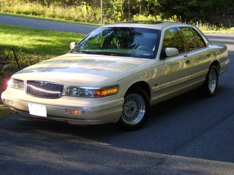 1996 Mercury Grand Marquis - A tan/champagne Mercury Grand Marquis four-door sedan photographed from the front-left 3/4 angle, parked on an asphalt driveway with grass…