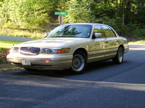 1996 Mercury Grand Marquis - A gold/champagne Mercury Grand Marquis full-size sedan parked at an angle on a paved road near a wooded intersection, shown from the front-…