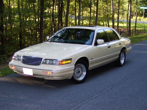 1996 Mercury Grand Marquis - A cream/beige four-door Mercury Grand Marquis sedan photographed from the front-left three-quarter angle, parked on an asphalt road with a…