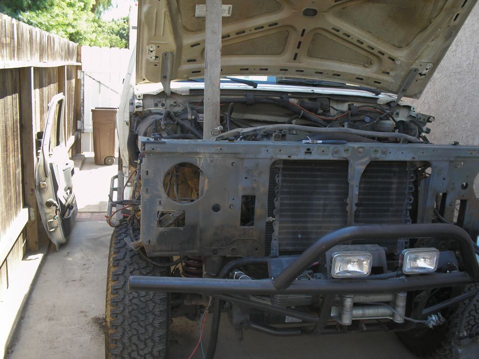 Front of a disassembled Ford Bronco with hood open, showing engine bay and radiator. Door is off and leaning against a fence