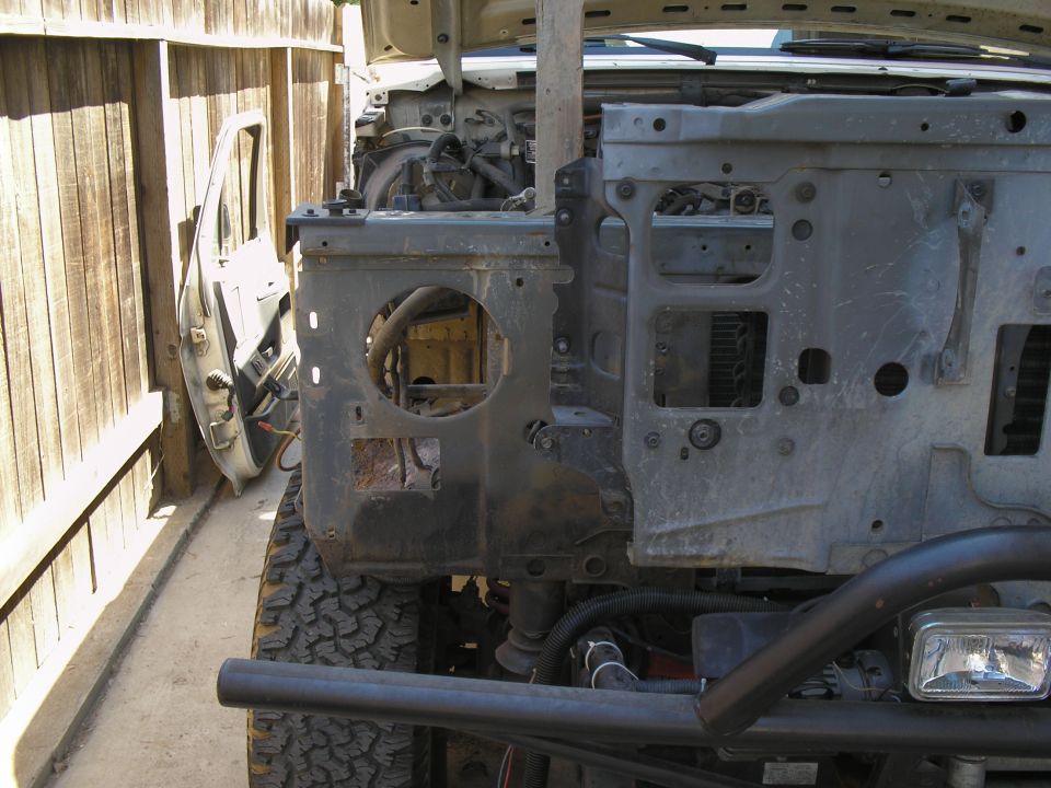 Disassembled vehicle front end with exposed engine components and a detached door leaning against a fence