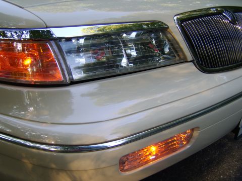 1996 Mercury Grand Marquis - Close-up of the front driver-side corner of a light gold/champagne sedan, showing the clear headlight assembly, amber corner turn signal, c…