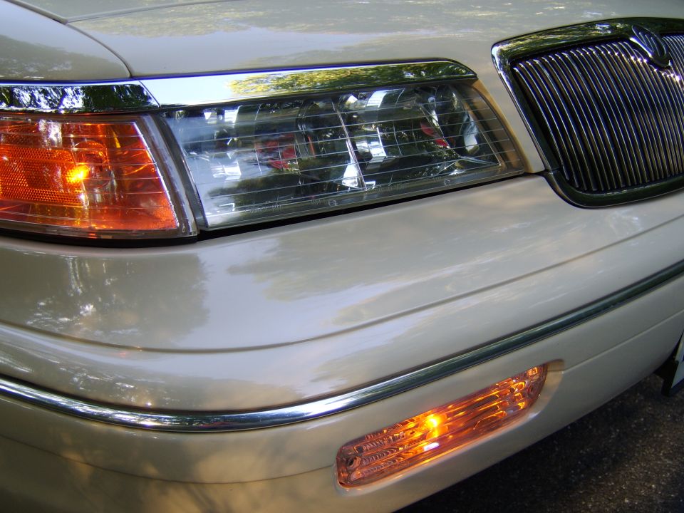 Front corner of gold sedan with headlight, chrome grille, and lit amber turn signals