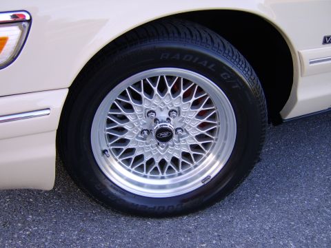 1996 Mercury Grand Marquis - Close-up of a cream/beige car's front wheel featuring a silver multi-spoke lacy mesh alloy wheel with a Ford center cap, mounted on a Coope…