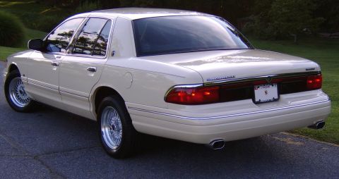 1996 Mercury Grand Marquis - A cream/white Mercury Grand Marquis full-size sedan photographed from the rear three-quarter angle, showing chrome trim, wire-spoke wheels,…