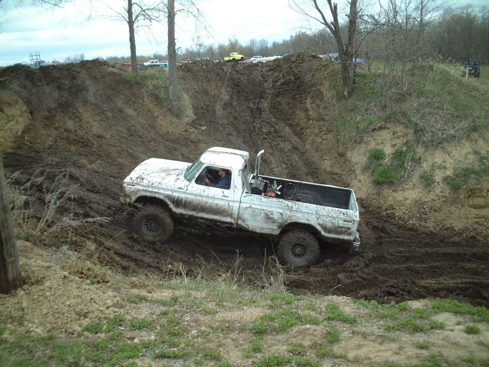 Muddy white lifted pickup truck climbing a steep dirt hill at an off-road mud bogging area