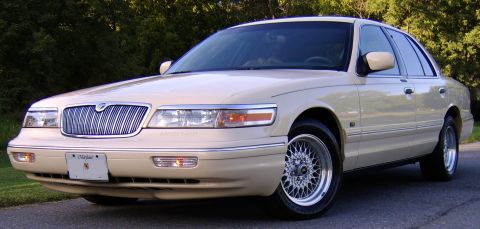 1996 Mercury Grand Marquis - A cream/beige Mercury Grand Marquis full-size sedan photographed from the front three-quarter angle, showing its chrome waterfall grille, d…