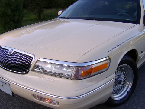 1996 Mercury Grand Marquis - Close-up 3/4 front view of a cream/beige Mercury sedan showing the chrome waterfall grille with Mercury emblem, clear headlight with amber…