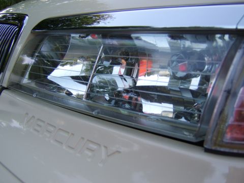 1996 Mercury Grand Marquis - Close-up of the passenger-side front headlight assembly on a light tan/beige Mercury, with the embossed 'MERCURY' lettering visible on the…