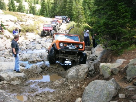 1995 Ford Bronco - A line of off-road vehicles, led by an orange first-generation Ford Bronco, navigates a challenging rocky stream bed.