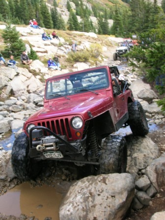 1995 Ford Bronco - A red Jeep Wrangler tackles a challenging off-road course, with other vehicles and spectators in the background.