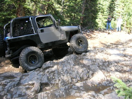 1995 Ford Bronco - A heavily modified Jeep Wrangler navigates a rocky off-road trail with two people watching in the background.