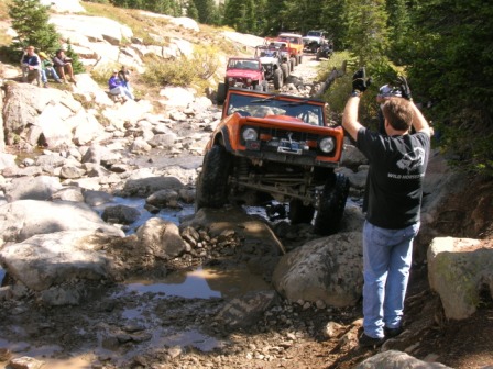 1995 Ford Bronco - Off-road vehicles navigate a challenging rocky stream on a trail, with spectators observing the side.